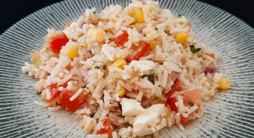 Une généreuse portion de salade de riz thon, garnie de dés de tomates, de maïs et d'œufs durs, sur une assiette texturée.
