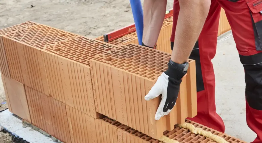 Murowanie ścian z porothermu. Budowlaniec w rękawicy zakłada kolejny pustak na pianę montażową.