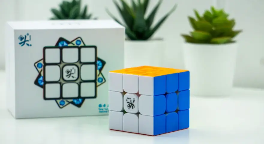 En speed cube 3x3 med orange, vit och blå sida står bredvid sin förpackning. Tre gröna växter i vita krukor syns i bakgrunden.