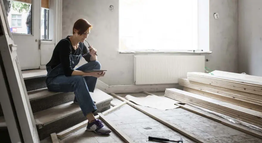 Femme en salopette assise sur des marches, tablette à la main, réfléchissant aux travaux de second œuvre. Matériaux de construction et outils au sol.