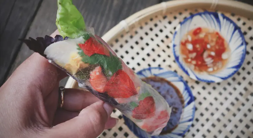 Ręka trzyma świeże rolls z papieru ryżowego z truskawkami i ziołami. Obok miseczki z sosem.