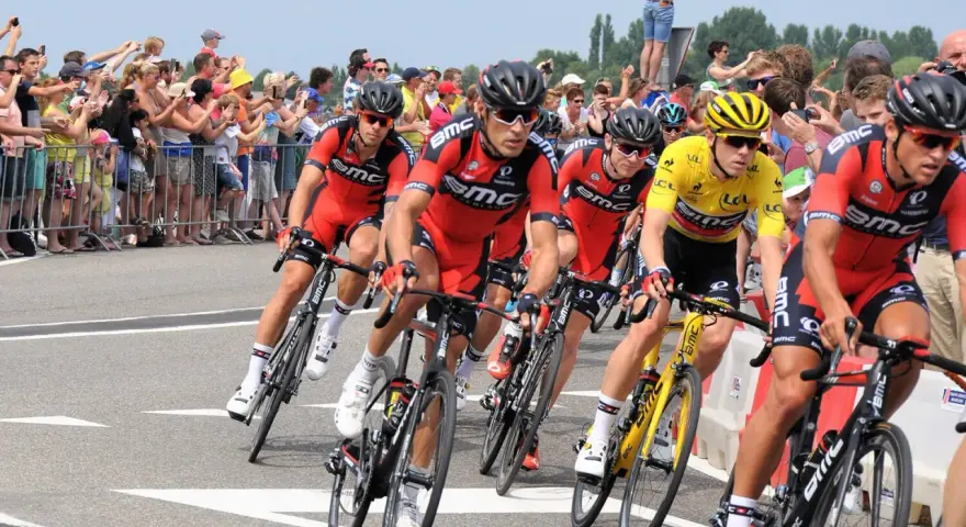 Cyclists in red BMC jerseys race, with one in yellow, as spectators cheer. A perfect scene to watch Tour de France live!
