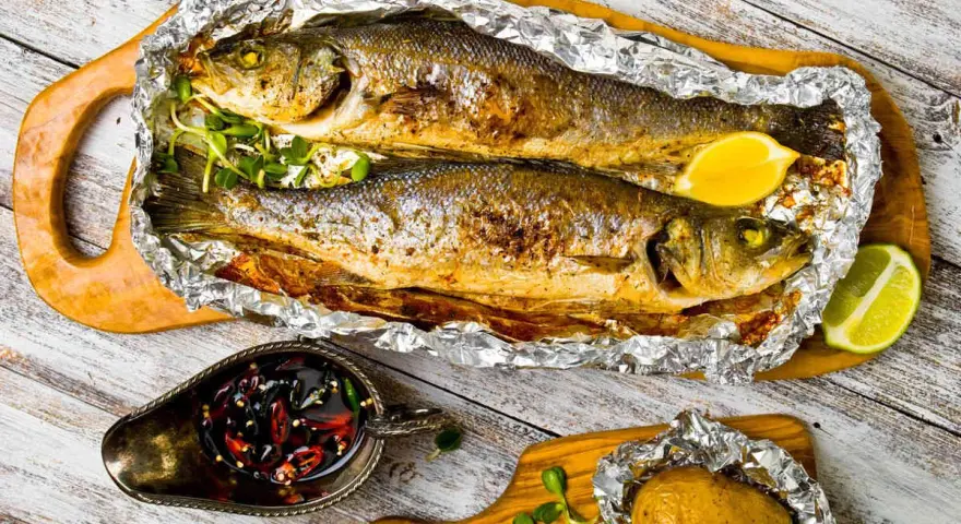 Due branzini cotti al cartoccio, dorati e succulenti, serviti con spicchi di limone, patata al cartoccio e salsa piccante.