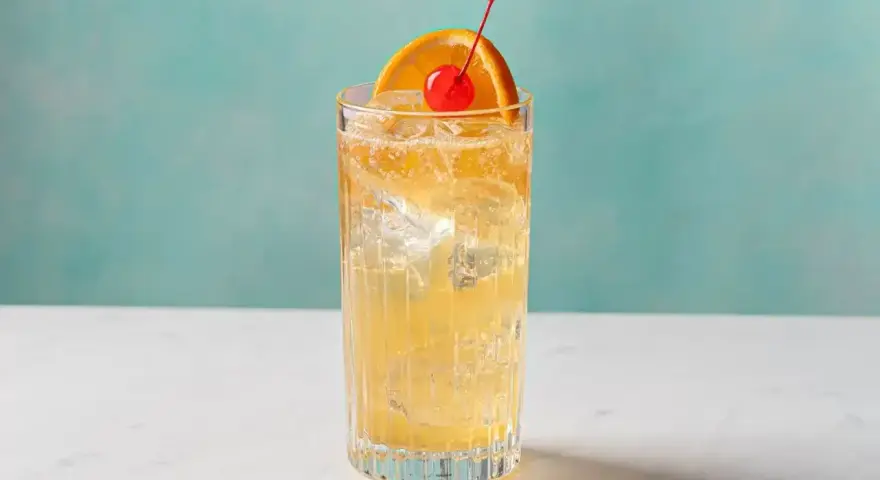 Orzeźwiający John Collins drink z lodem, plasterkiem pomarańczy i wisienką na tle turkusowej ściany.