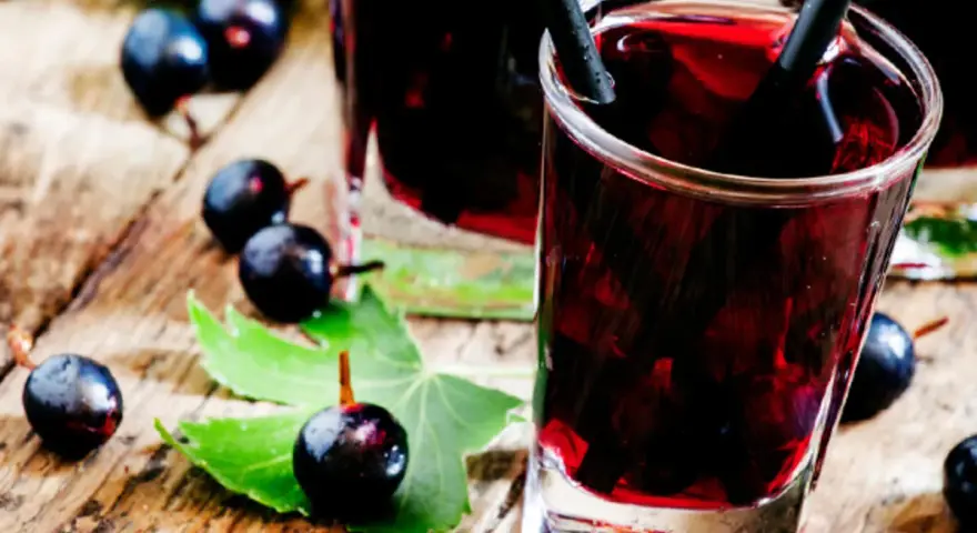 Gläser mit dunklem Johannisbeeren Schnaps, umgeben von frischen Beeren und Blättern auf Holz.