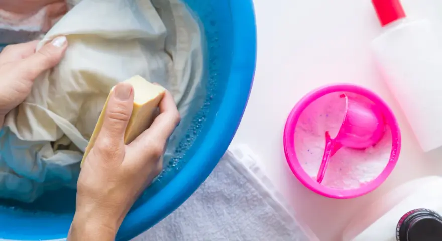 Ręczne pranie poplamionego ubrania w niebieskiej misce z wodą i mydłem. Obok proszek do prania i butelki detergentów. Jak sprać farbę do włosów z ubrań.