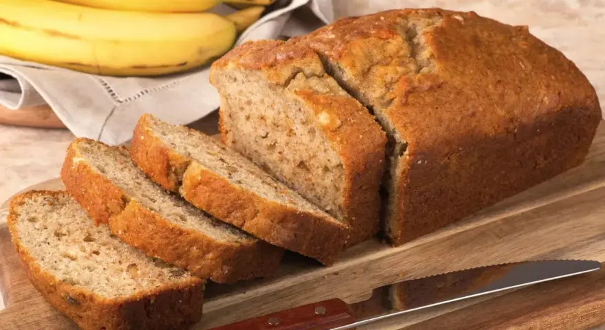 Pyszny, domowy chlebek bananowy, pokrojony na kromki, leży obok dojrzałych bananów na desce.