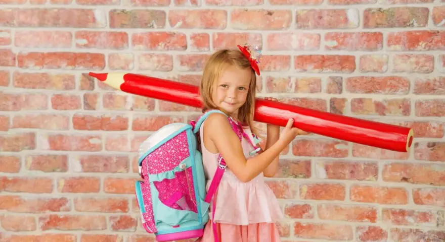 Ein Mädchen mit pinkem Rucksack und riesigem roten Stift vor Ziegelwand. Bereit für die Einschulung Berlin 2025!
