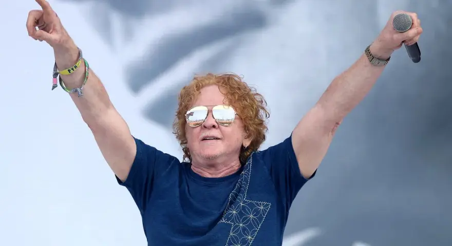 Wokalista Simply Red z uniesionymi rękami, trzymający mikrofon. Ma charakterystyczne rude włosy i okulary przeciwsłoneczne.
