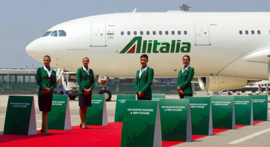 Cztery stewardessy w zielonych uniformach stoją na czerwonym dywanie przed samolotem Alitalia.