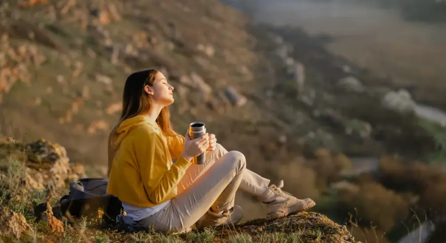 A young woman enjoys the art of doing nothing, holding a thermos and basking in the sun on a rocky hillside.
