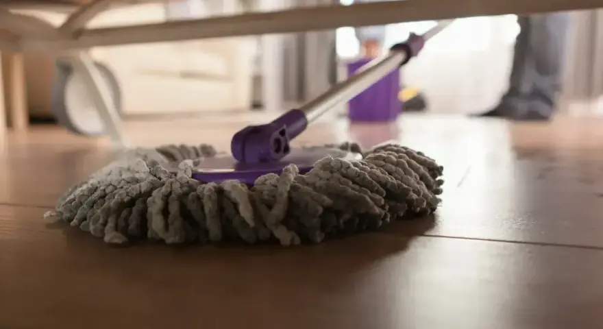 Mop pod łóżkiem, gotowy do czyszczenia paneli. W tle widać wiadro i nogi osoby sprzątającej.