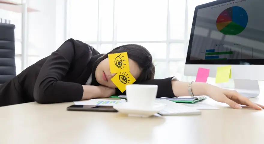 En kvinna sover vid sitt skrivbord med post-it-lappar som föreställer ögon på ögonlocken. Det är uppenbart att hon tycker det är tråkigt på jobbet.