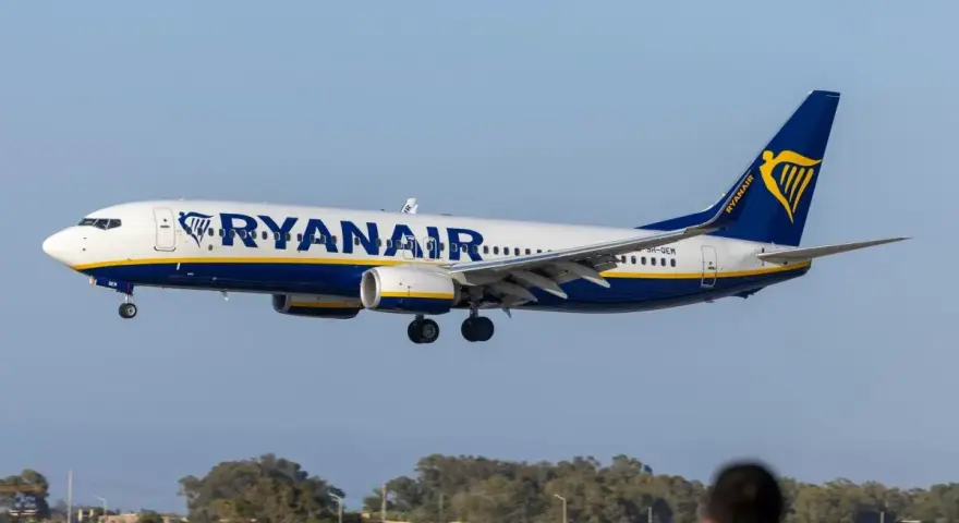 Samolot Ryanair lądujący na tle nieba i drzew, z widoczną sylwetką osoby w pierwszym planie.