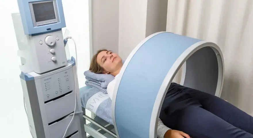 Kobieta leży na łóżku rehabilitacyjnym, poddając się zabiegowi z wykorzystaniem urządzenia generującego pole magnetyczne, które obejmuje jej kolano.