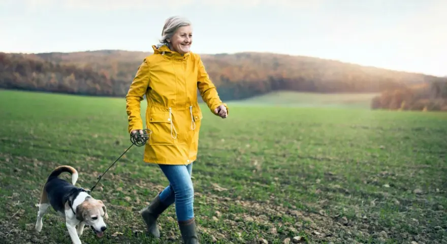 Glad kvinna i gul jacka njuter av en promenad med sin hund i ett fält. En härlig promenad som bränner kalorier.