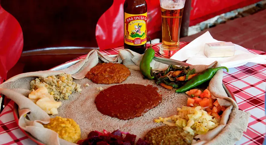 Tradycyjna kuchnia etiopska na płaskim chlebie injera, z różnymi gulaszami, warzywami i papryczkami. Obok piwo.