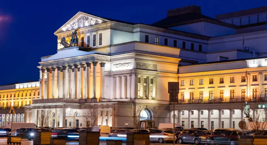 Teatr Wielki w Warszawie, najstarszy teatr w Polsce, nocą. Monumentalna fasada z kolumnami i rzeźbami, oświetlona ciepłym światłem, z samochodami w ruchu ulicznym przed budynkiem.