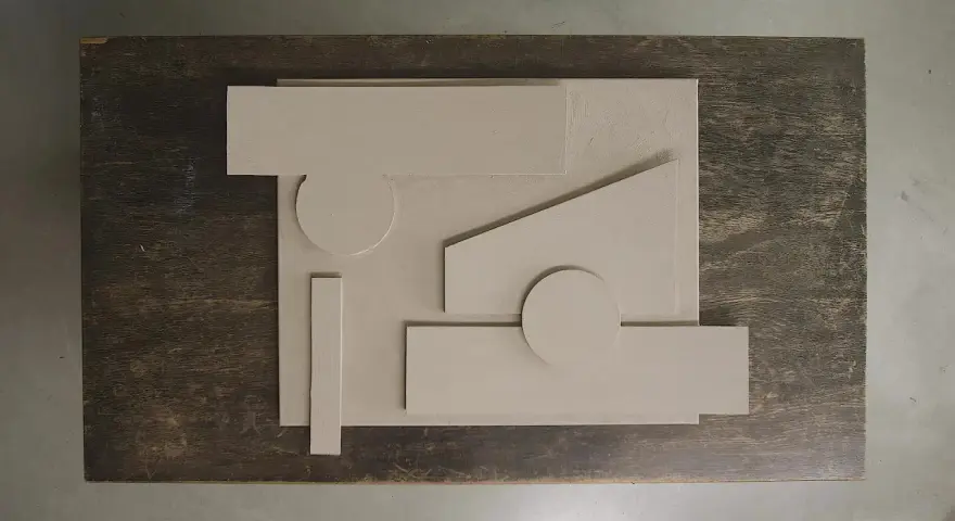 Abstrakt komposition med geometriska former i ljusbeige akrylfärg, arrangerade på en mörk träyta.