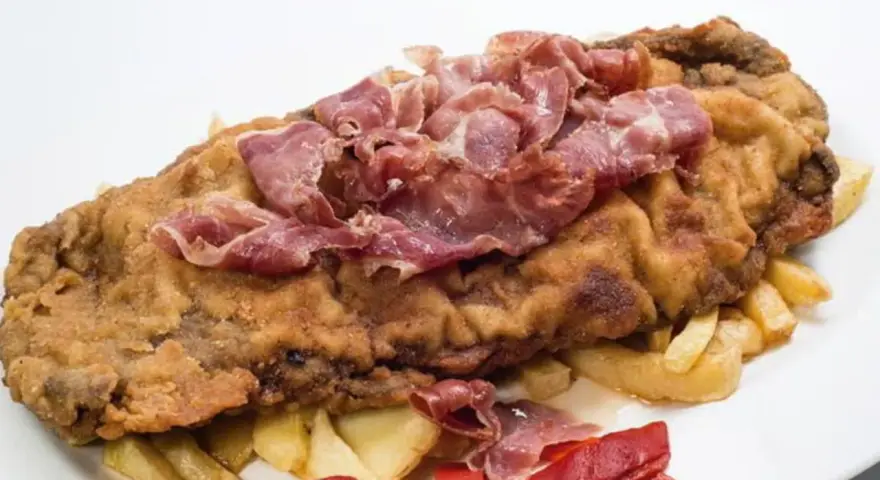 Cachopo asturiano rebozado con jamón serrano encima, acompañado de patatas fritas y pimientos rojos.