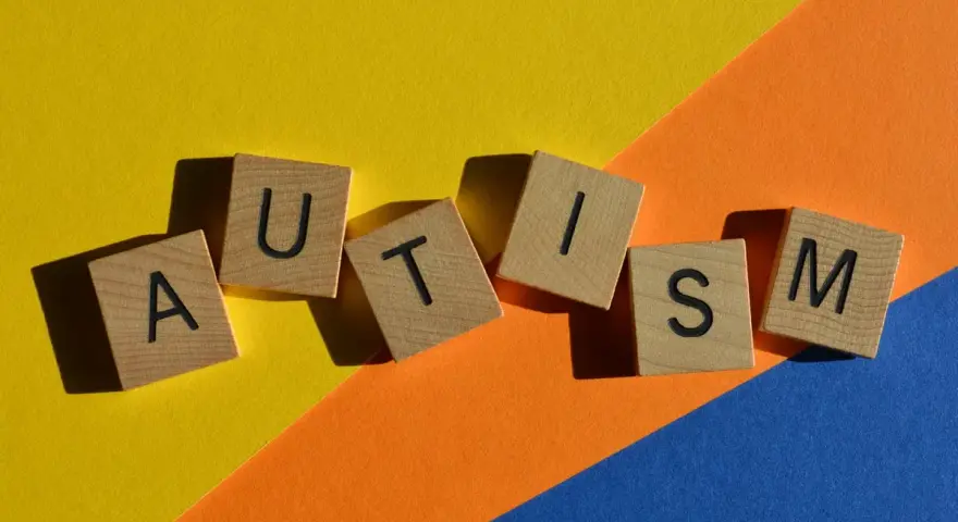 Lettere di legno formano la parola AUTISM su uno sfondo colorato. La domanda "autistici si nasce o si diventa" risuona.