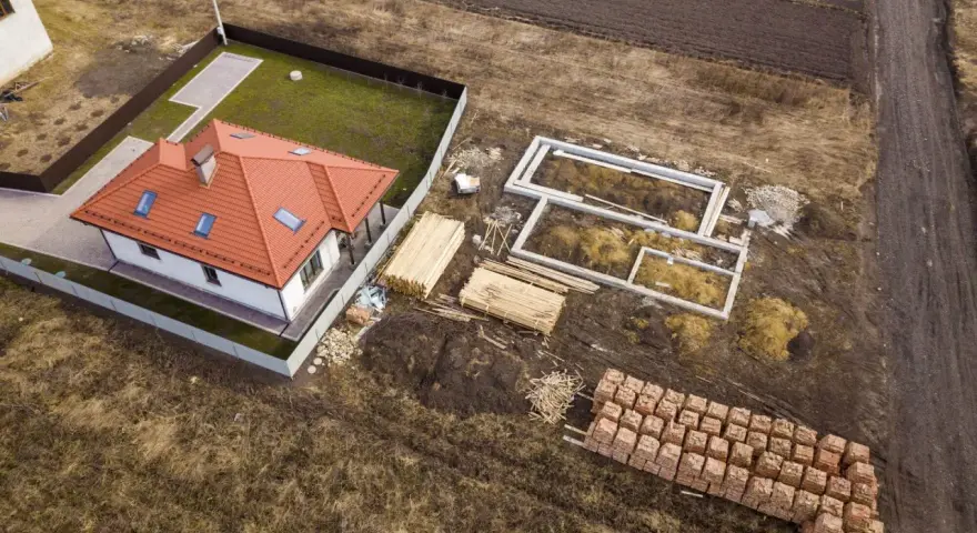 Zabudowa zagrodowa w budowie: gotowy dom z czerwonym dachem, fundamenty, drewno i palety cegieł na placu budowy.