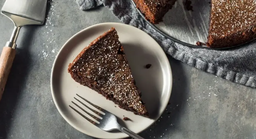 Una fetta di torta all'acqua al cacao, spolverata di zucchero a velo, su un piatto con una forchetta. Accanto, la torta intera e una paletta.