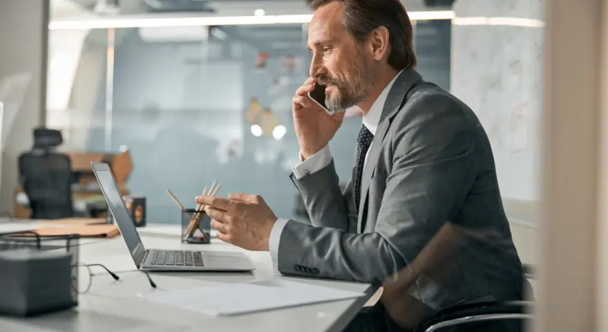 Biznesmen w garniturze rozmawia przez telefon, pracując przy laptopie. To typowy dzień w życiu CEO, co to znaczy być liderem.