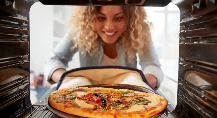 Uśmiechnięta kobieta wyjmuje z piekarnika pyszną, gorącą pizzę. Właśnie skończyła piec pizzę i jest gotowa do jedzenia.