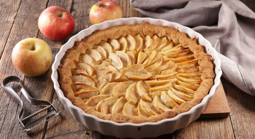 Torta di mele umida e soffice, con mele fresche e un pelapatate su un tavolo di legno rustico.