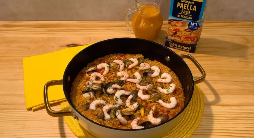 Delicioso arroz a la marinera en paellera, con gambas y mejillones. Al lado, un cartón de caldo para paella de marisco y una jarra con líquido naranja.
