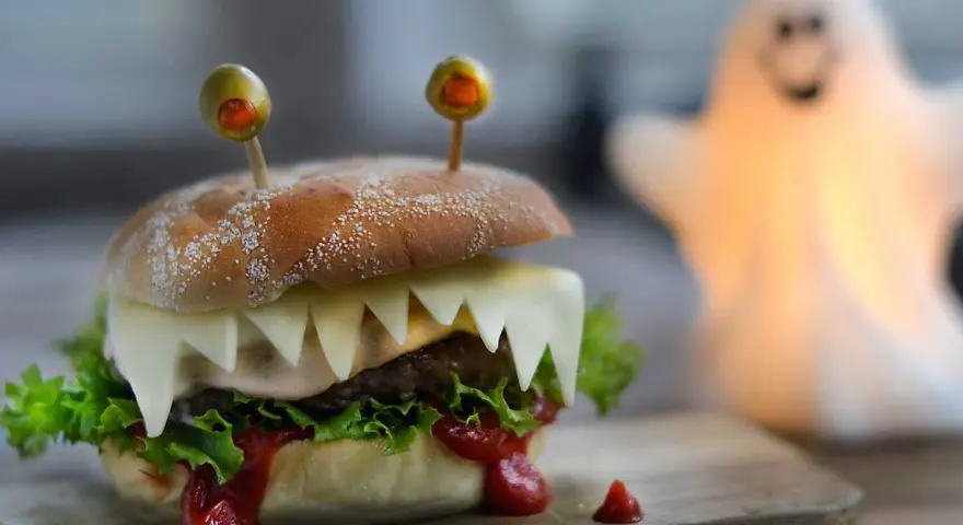 En läskig halloween hamburgare med osttänder och olivögon. En spöklampa lyser upp i bakgrunden.