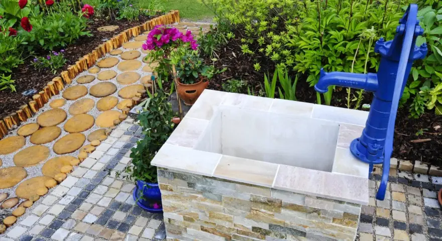Ein Gartenweg führt zu einem frisch gebauten Brunnen mit blauer Handpumpe.