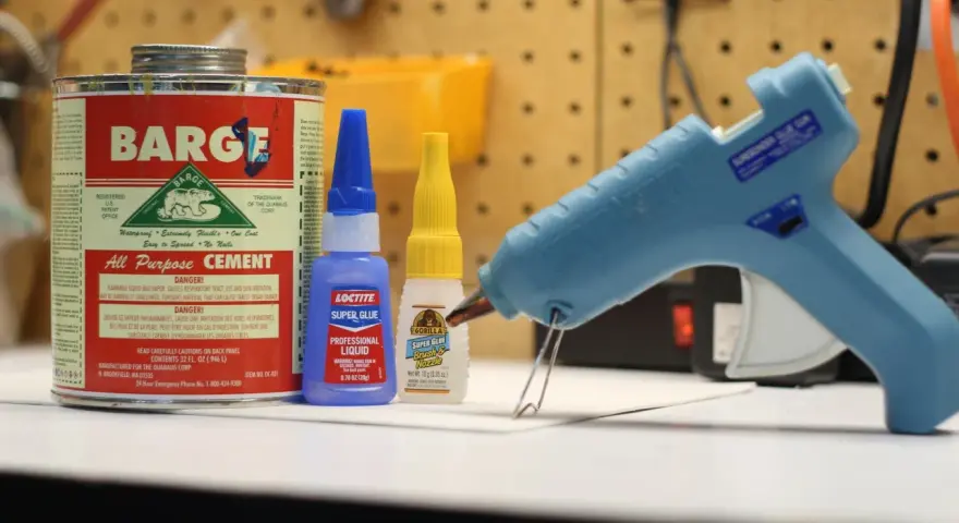 A hot glue gun, Barge cement, Loctite super glue, and Gorilla Super Glue are on a workbench. Does Gorilla Glue melt? This image shows various adhesives.