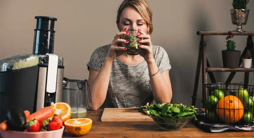 Kvinna dricker grön juice, omgiven av färsk frukt och grönsaker. Hon verkar njuta av sin hälsosamma dryck, kanske som en del av hur deffar man.