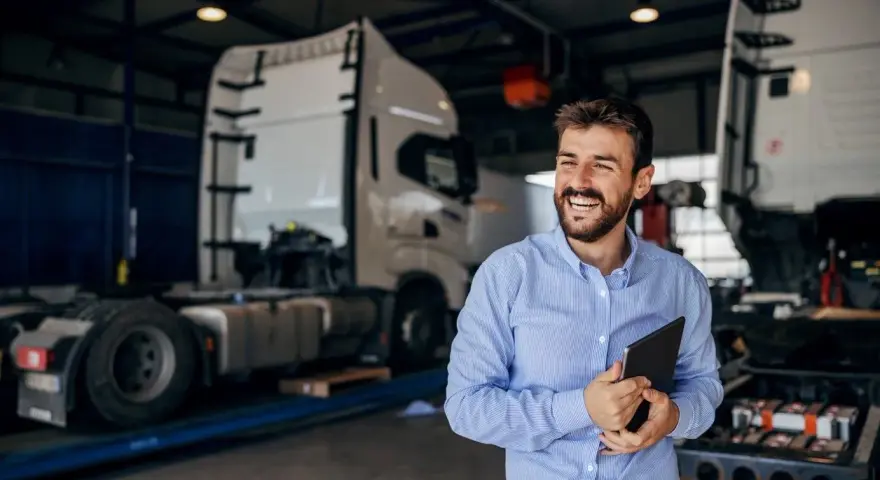 Uśmiechnięty mężczyzna z tabletem w ręku, w tle warsztat z ciężarówkami. Oferuje **usługę transportową ryczałt**.