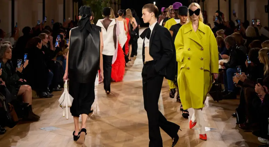 Sfilata Balenciaga: modelli con abiti audaci, tra cui un cappotto giallo acceso e un completo nero, sotto gli occhi del pubblico.