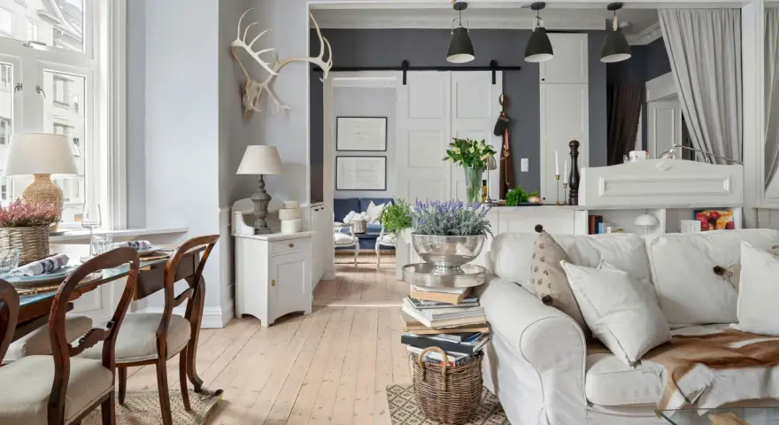 Un salotto con un'atmosfera da arredamento francese chic. Tavolo da pranzo in legno, divano bianco accogliente, lampade e decorazioni rustiche.