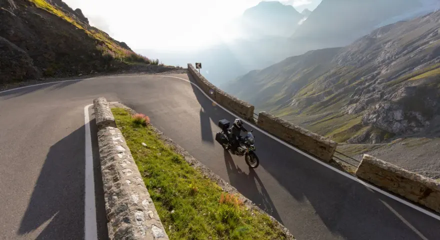 Motocyklista przemierza serpentyny górskiej drogi, odkrywając najpiękniejsze trasy motocyklowe w Europie. Słońce przebija się przez mgłę, oświetlając majestatyczne szczyty.