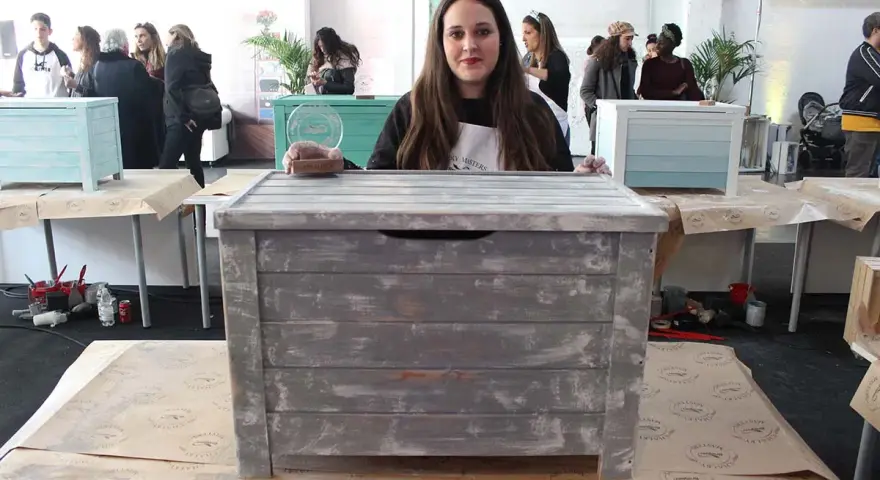 Una mujer muestra un baúl pintado a la tiza con un acabado desgastado. Otros baúles decorados se ven en el fondo.