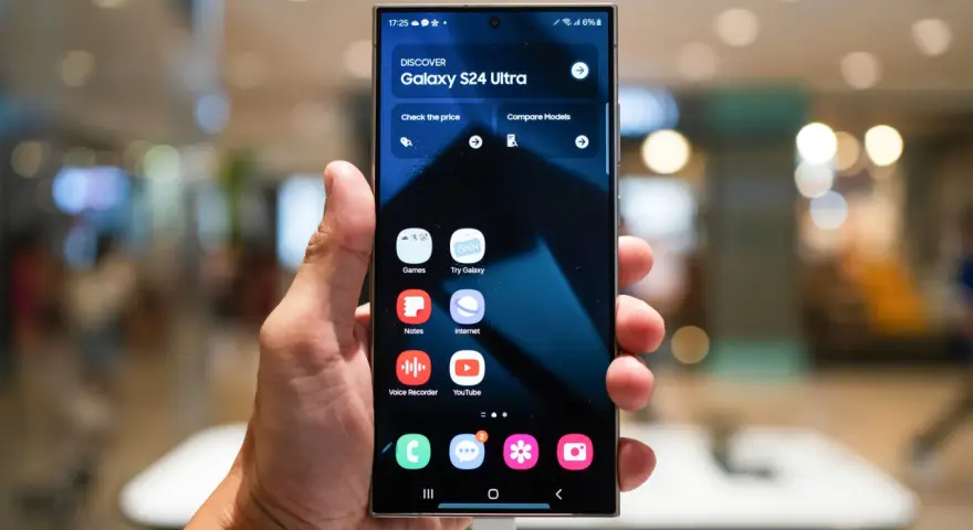 Dłoń trzyma smartfon z interfejsem One UI 6, prezentującym ekran startowy Galaxy S24 Ultra z ikonami aplikacji.