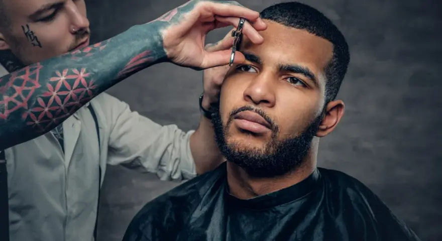 Barbero con tatuajes en el brazo recorta las cejas de un hombre con barba, creando un look buzz cut.