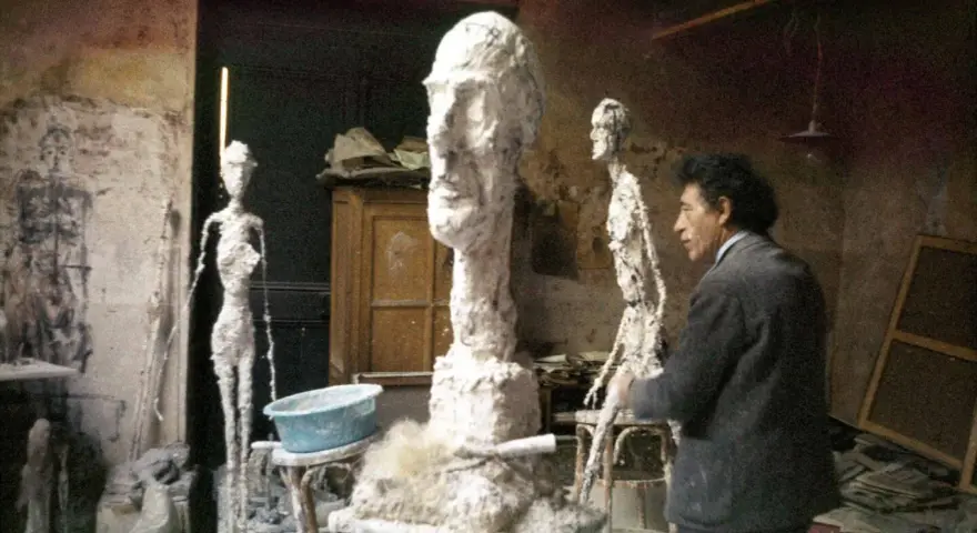 Alberto Giacometti w pracowni, otoczony swoimi charakterystycznymi, smukłymi rzeźbami i niedokończonymi formami.