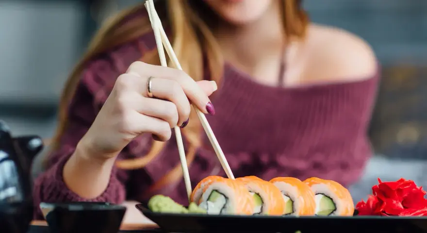 Uśmiechnięta kobieta w ciąży sięga po sushi pałeczkami. Na talerzu łosoś, awokado i imbir.