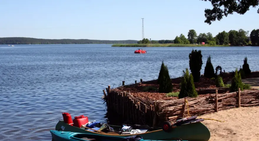 Dwie kajaki na piaszczystej plaży w Swornegaciach na Kaszubach. W oddali czerwona łódka i jezioro.