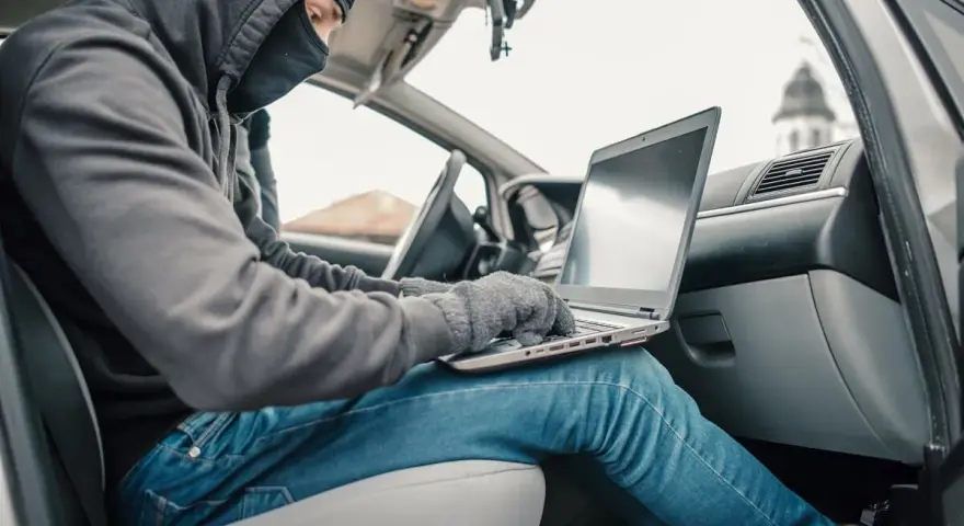 Haker w masce i kapturze próbuje obejść zabezpieczenia keyless go, używając laptopa w samochodzie.