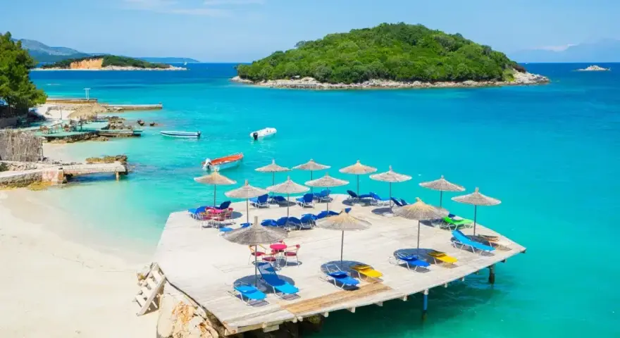 Rajska plaża w Albanii, turkusowe morze, drewniany pomost z leżakami i parasolami, zielona wyspa w oddali.