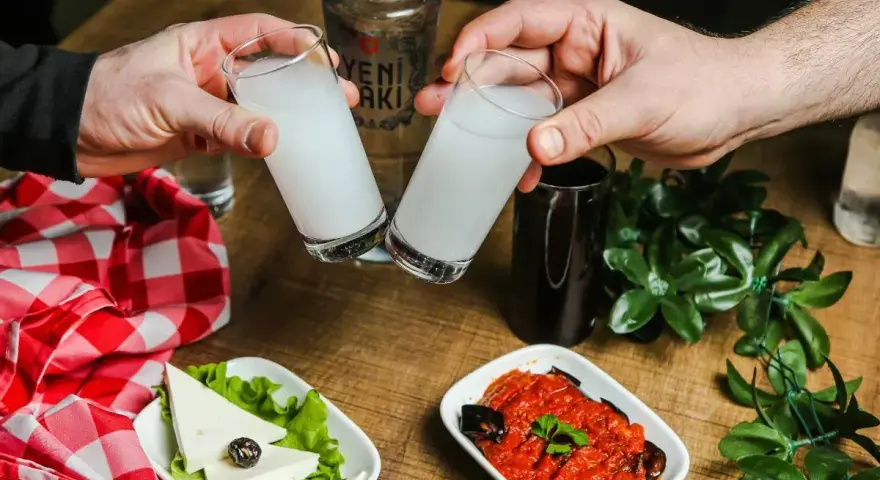 Dwie szklanki z rakı, tradycyjnym tureckim alkoholem, unoszą się do toastu. Obok ser, sałata, sos pomidorowy z bakłażanem i butelka rakı. Idealne połączenie na wieczór.