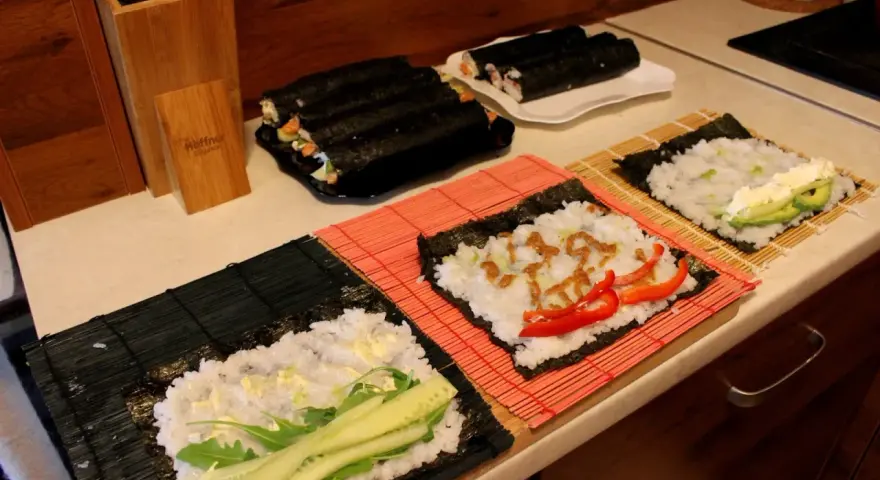 Przygotowanie ryżu do sushi. Na matach leżą arkusze nori z ryżem i dodatkami: ogórkiem, awokado, papryką i sosem.