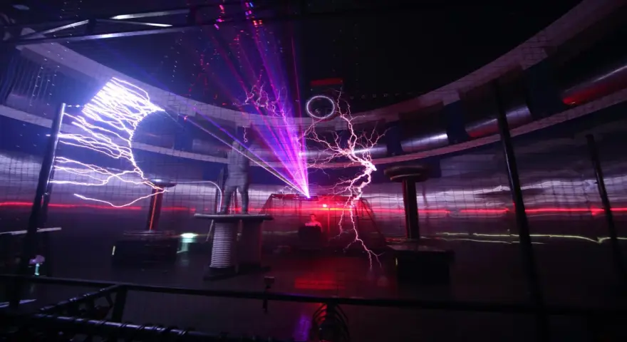 Spektakuläre Blitze zucken in einem **Faradayschen Käfig**, während ein Mann sicher darin steht. Laserstrahlen durchschneiden die Dunkelheit.